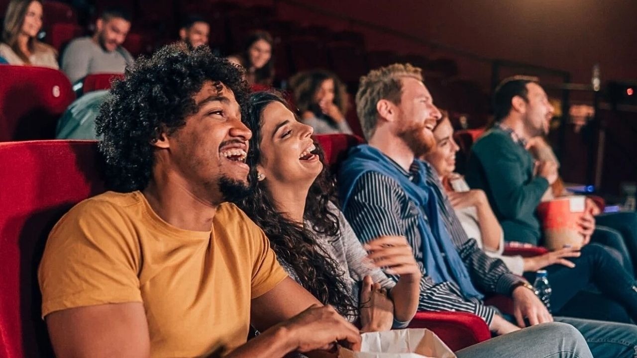 Pessoas diversas riem em sala de cinema, com assentos vermelhos e pipoca.