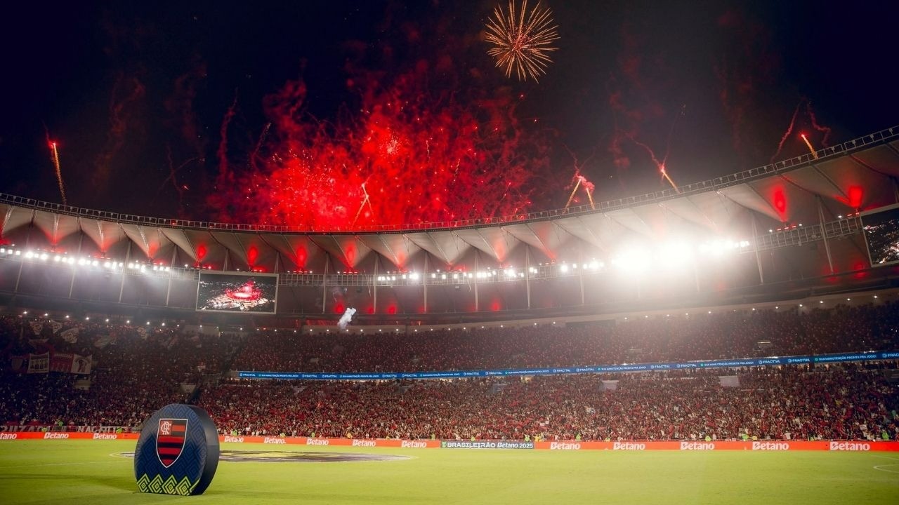 Maracanã lotado, fogos vermelhos no céu noturno, objeto com escudo do Flamengo no gramado.