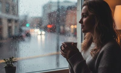 Mulher em perfil olhando a chuva pela janela, segurando caneca fumegante, luzes desfocadas na rua.