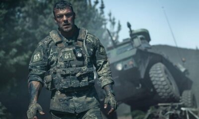 Homem em uniforme militar camuflado, colete tático e luvas, com veículo blindado em segundo plano.