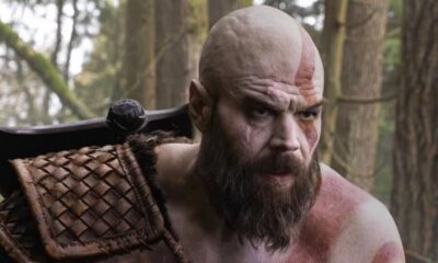 Homem careca barbudo com marca vermelha no rosto e corpo, armadura de couro, em floresta.