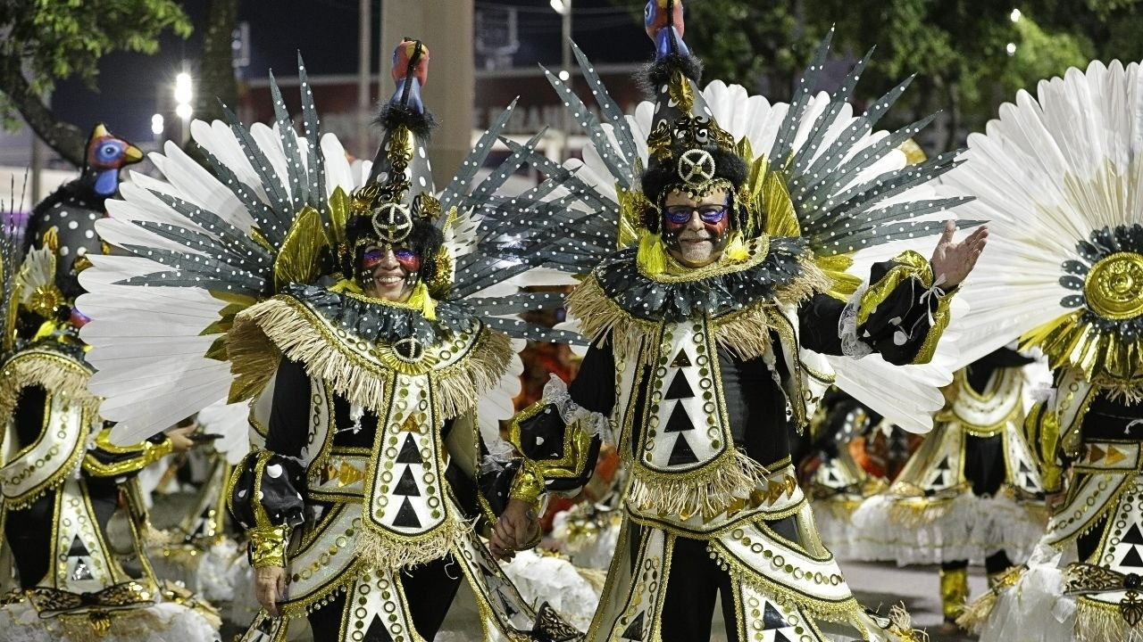 Duas pessoas sorridentes em fantasias pretas, brancas e douradas de Carnaval com pintura facial.