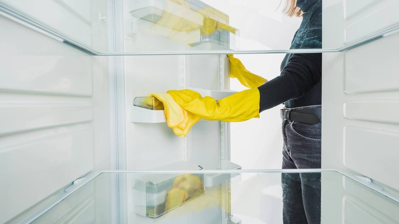 Pessoa com luvas amarelas e pano amarelo limpando o interior branco de uma geladeira com prateleiras de vidro.