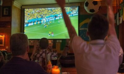 Pessoas assistem jogo de futebol em telão de bar escuro; bandeira do Brasil ao lado.