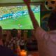Pessoas assistem jogo de futebol em telão de bar escuro; bandeira do Brasil ao lado.