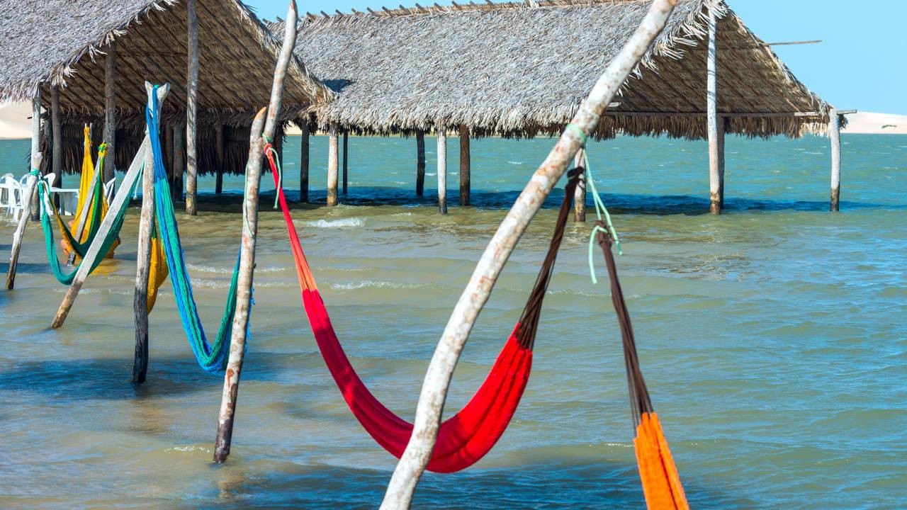 Redes coloridas suspensas sobre a água em um lago, com cabanas de palha no fundo.