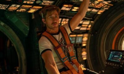 Ryan Gosling em um cockpit de nave espacial, com cinto de segurança laranja e óculos.