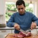 Um homem de suéter azul corta carne crua com faca e pinça em uma tábua de madeira na cozinha.