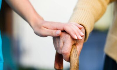 Mão jovem tocando mão envelhecida que segura uma bengala de madeira.