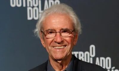 Juca de Oliveira sorrindo, óculos, cabelos grisalhos, em close-up. Fundo escuro.