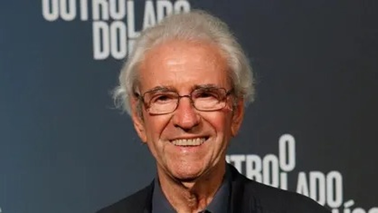 Juca de Oliveira sorrindo, óculos, cabelos grisalhos, em close-up. Fundo escuro.