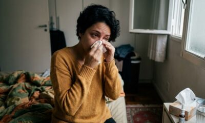 Mulher com lenço no nariz, sentada em quarto com caixa de lenços e spray nasal.