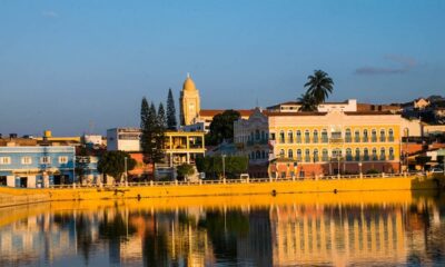 Cidade colorida refletida em lago calmo sob céu azul, com arquitetura histórica e vegetação.