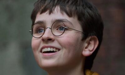 Menino jovem de cabelo castanho, óculos redondos e olhos azuis sorrindo para cima.