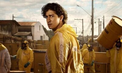 Homem de cabelo cacheado em traje amarelo escrito MÁRCIO, olhando para trás, com outros em trajes e barris.