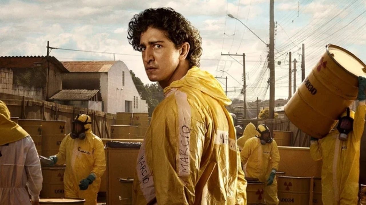 Homem de cabelo cacheado em traje amarelo escrito MÁRCIO, olhando para trás, com outros em trajes e barris.