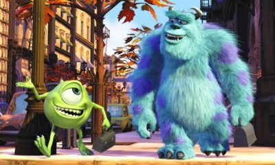 Monstros Mike Wazowski e Sulley sorrindo em uma calçada de cidade fictícia, com prédios ao fundo.