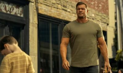 Ator Alan Ritchson, musculoso, camiseta verde, caminha em rua urbana; um menino abaixado na frente.