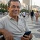 Homem sorridente de meia-idade segura smartphone sentado em banco no calçadão do Rio.