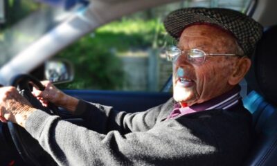 Homem idoso ao volante de um carro, usando boné e óculos.