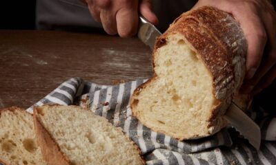 Mãos fatiam pão rústico de casca dourada e miolo claro sobre pano listrado em madeira.