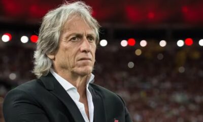 Jorge Jesus, técnico português, em close-up, vestindo blazer preto com fundo desfocado de estádio.