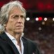 Jorge Jesus, técnico português, em close-up, vestindo blazer preto com fundo desfocado de estádio.