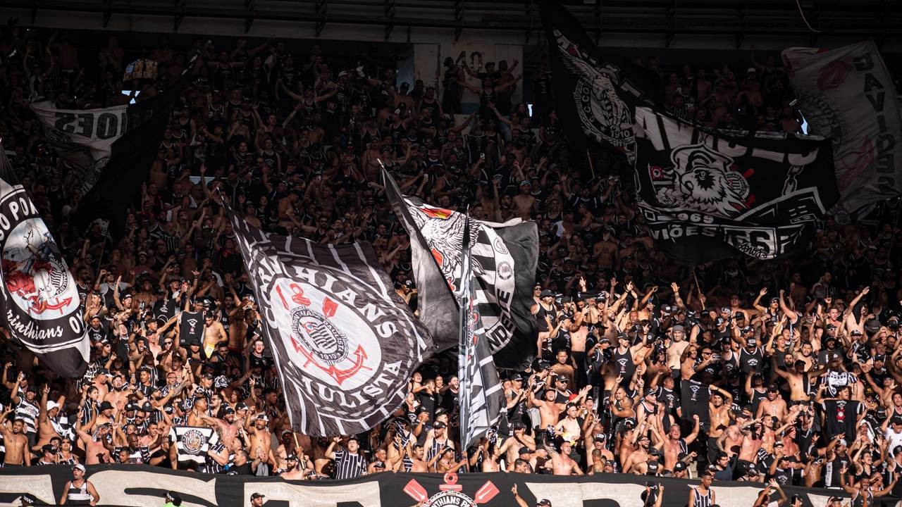 Multidão de torcedores do Corinthians em arquibancada, agitando bandeiras pretas e brancas do clube.
