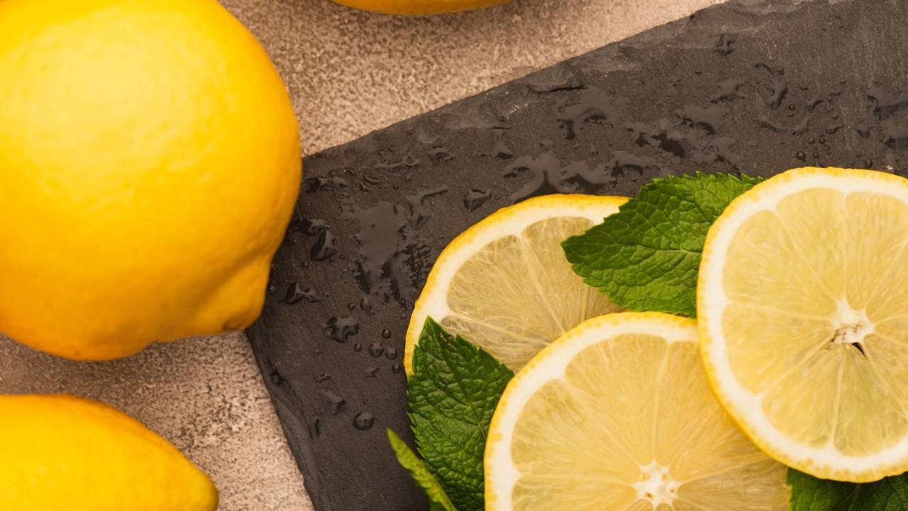 Dois limões inteiros, fatias de limão e folhas de hortelã sobre uma superfície escura molhada.