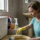 Mulher com luvas amarelas limpa vaso sanitário com escova, produtos de limpeza ao lado.