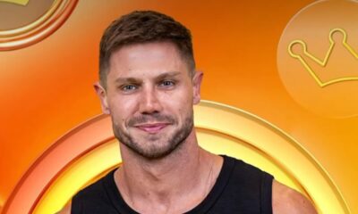 Homem branco, cabelo e barba castanhos, sorrindo levemente, camiseta preta, fundo laranja e coroa.