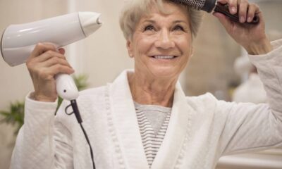 Mulher idosa sorrindo, secando cabelo grisalho com secador e escova, vestindo roupão branco.