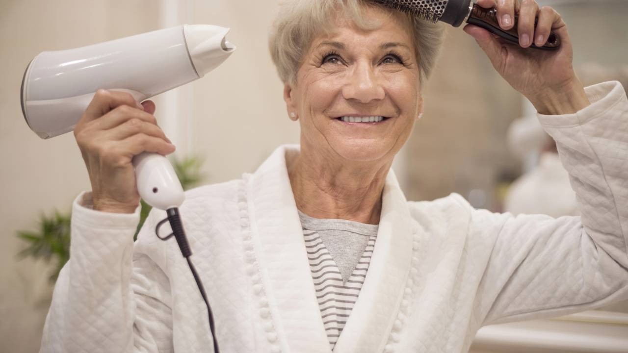Mulher idosa sorrindo, secando cabelo grisalho com secador e escova, vestindo roupão branco.