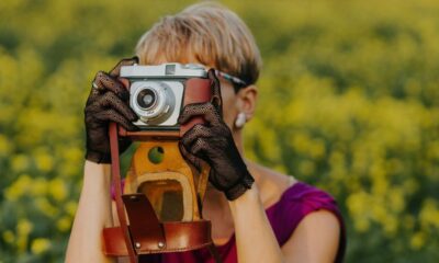 Pessoa de cabelos curtos segurando câmera analógica vintage com luvas pretas em campo amarelo.