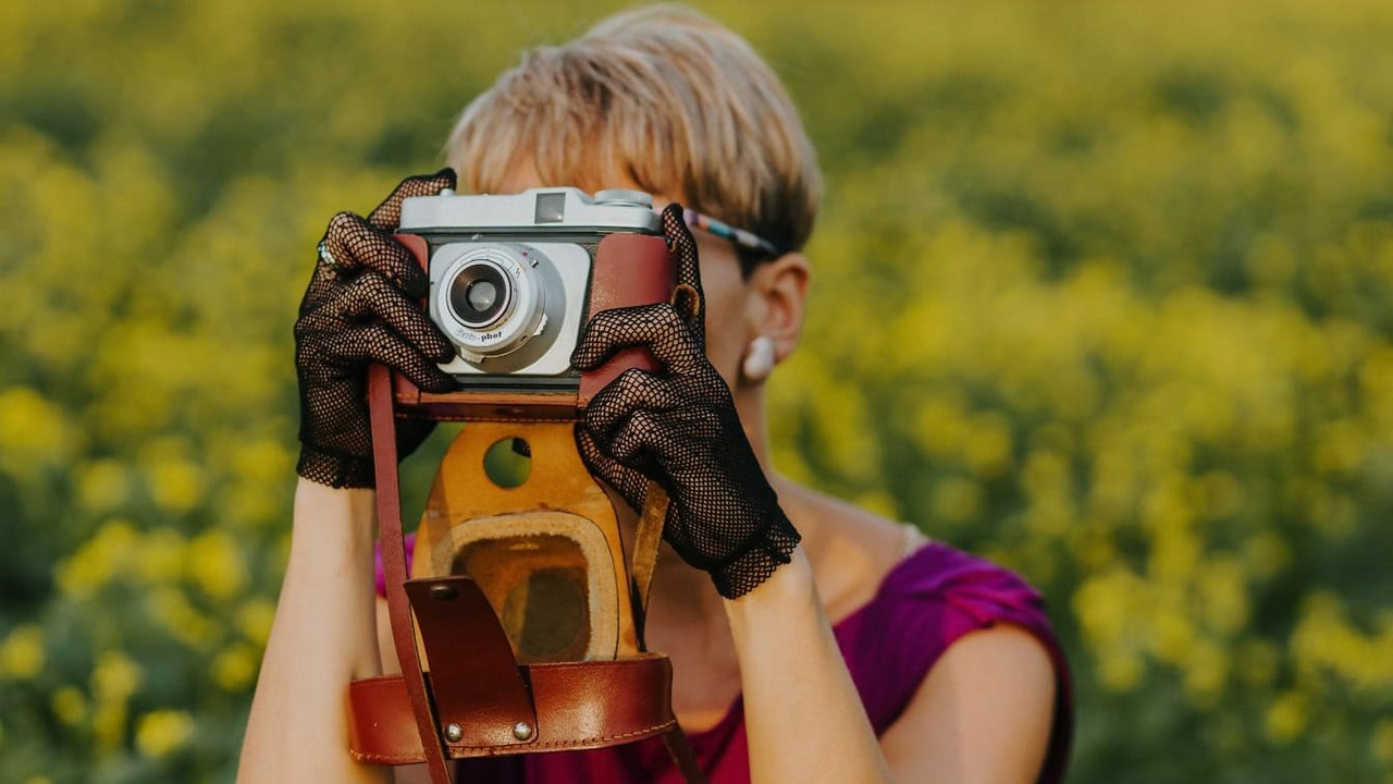 Pessoa de cabelos curtos segurando câmera analógica vintage com luvas pretas em campo amarelo.
