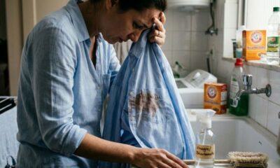 Mulher aflita segura camisa azul manchada. Ao fundo, pia com vinagre e bicarbonato de sódio.