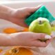Mãos usando uma esponja verde para lavar uma maçã verde com espuma em uma pia.