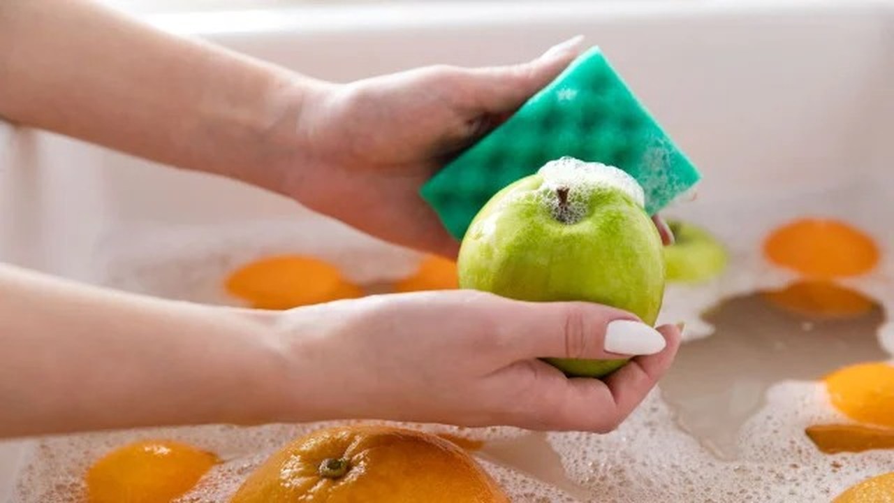 Mãos usando uma esponja verde para lavar uma maçã verde com espuma em uma pia.