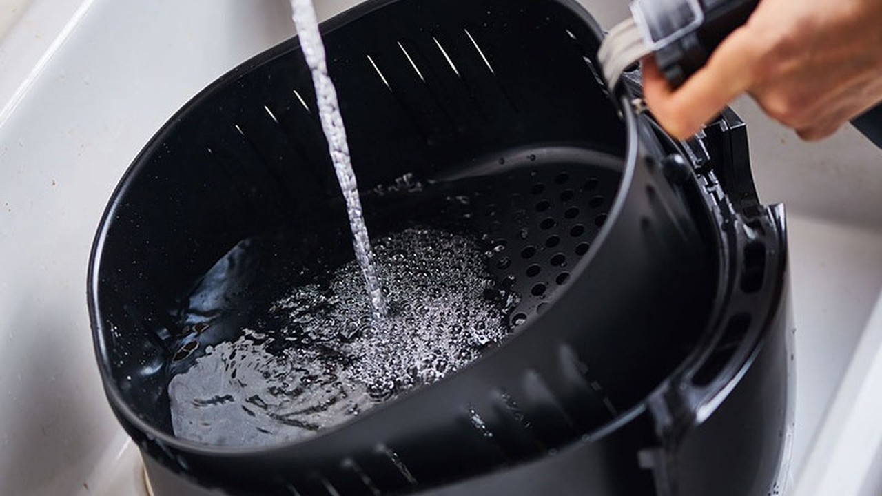Mão lavando cesto preto de air fryer com água corrente em pia branca.