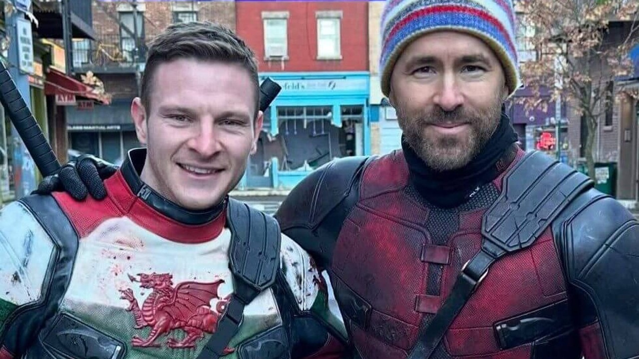 Ryan Reynolds barbudo com gorro e fantasia de Deadpool ao lado de homem sorrindo com camisa galesa.