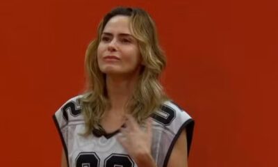 Ana Paula Renault em camiseta esportiva, tocando o peito com expressão emocionada, contra parede laranja.