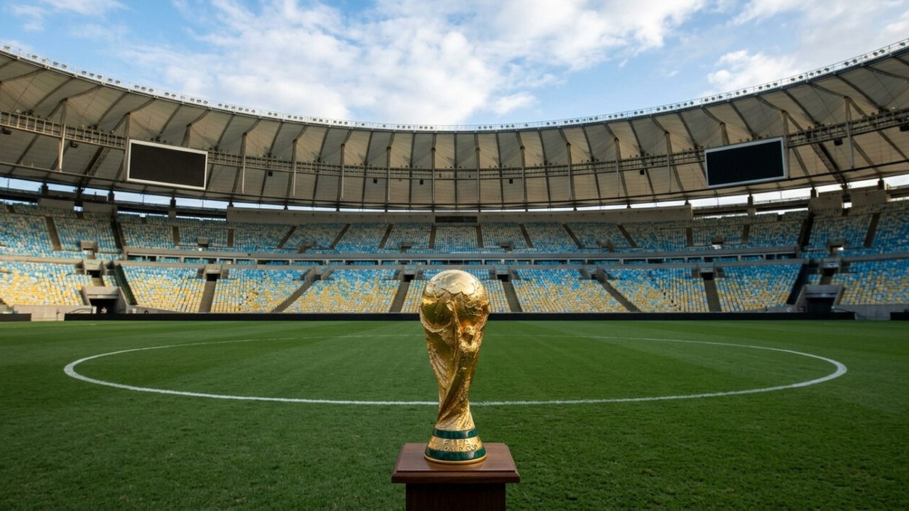 Troféu dourado da Copa do Mundo em campo de futebol vazio com arquibancadas azuis e amarelas.