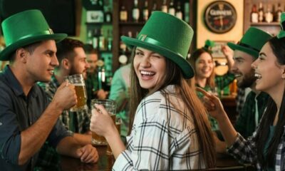 Jovens celebram no bar com chapéus e adereços verdes, segurando canecas de cerveja, sorrindo.