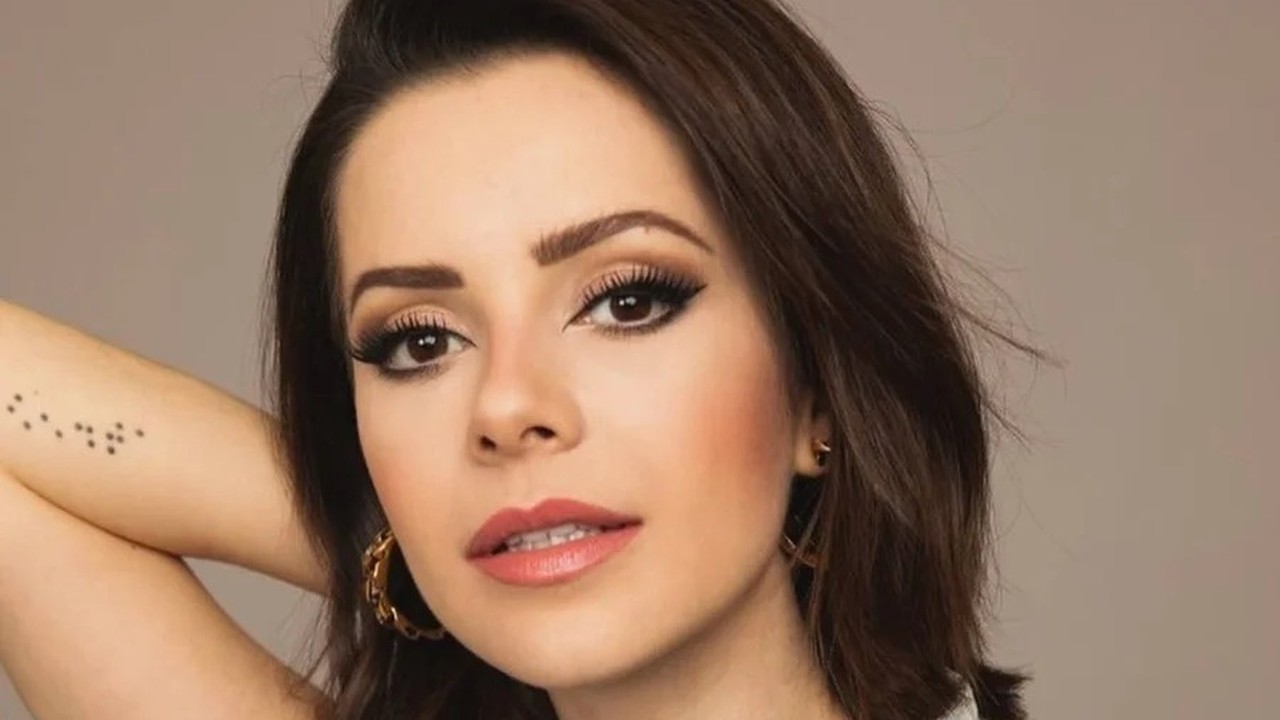 Close-up da cantora Sandy sorrindo, maquiagem, brincos dourados e tatuagem discreta no braço.