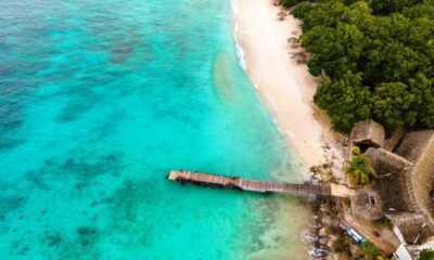 Vista aérea de praia caribenha: mar azul, areia branca, píer de madeira e telhados de palha.