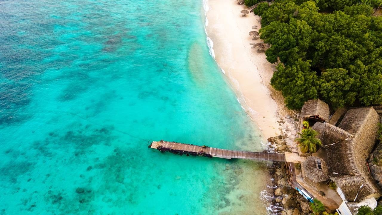 Vista aérea de praia caribenha: mar azul, areia branca, píer de madeira e telhados de palha.