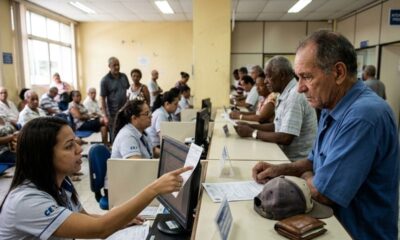 Mulher atendente em guichê aponta tela de computador para homem idoso, com outras pessoas aguardando.