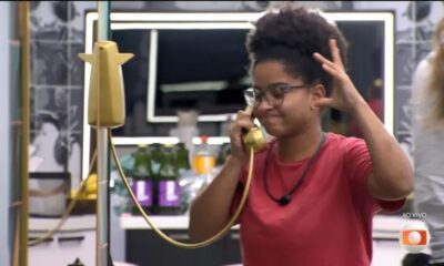 Mulher de óculos e cabelo crespo atende telefone dourado com expressão de leve sorriso.