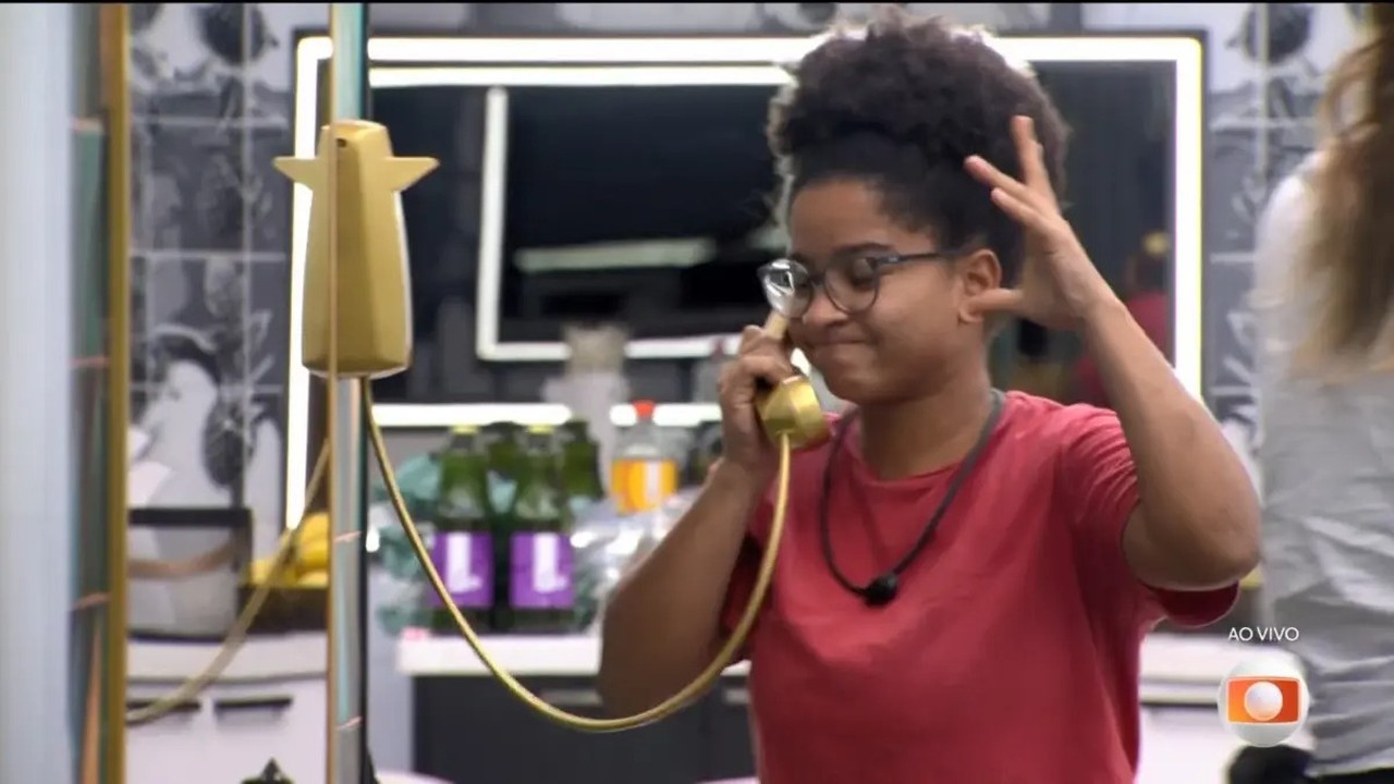 Mulher de óculos e cabelo crespo atende telefone dourado com expressão de leve sorriso.