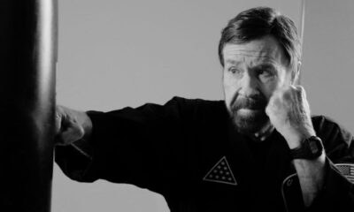 Homem idoso, Chuck Norris, com barba, soca saco de pancadas. Foto em preto e branco.